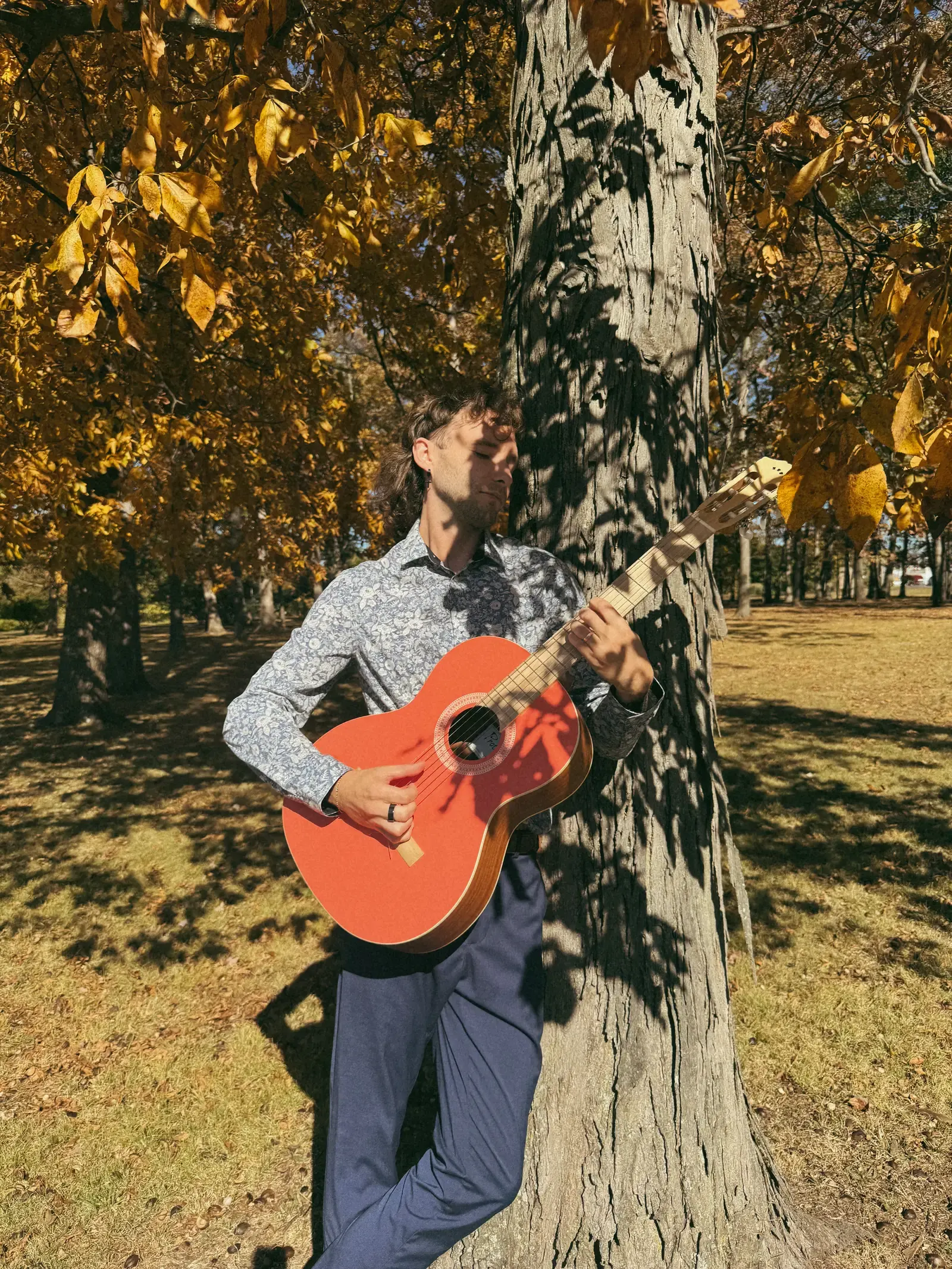 Mathew standing beneath a tree with his orange-red acoustic guitar.