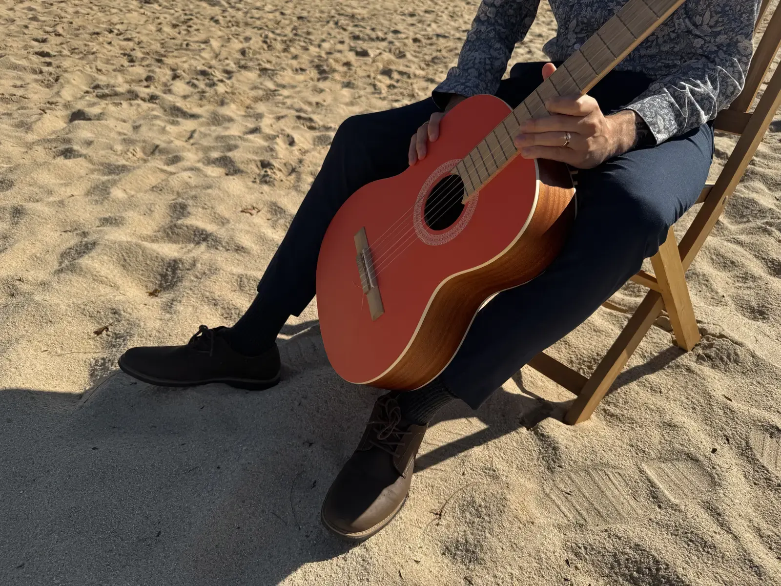 A close view of Mathew’s hands on his orange-red guitar, seated on a sandy path.