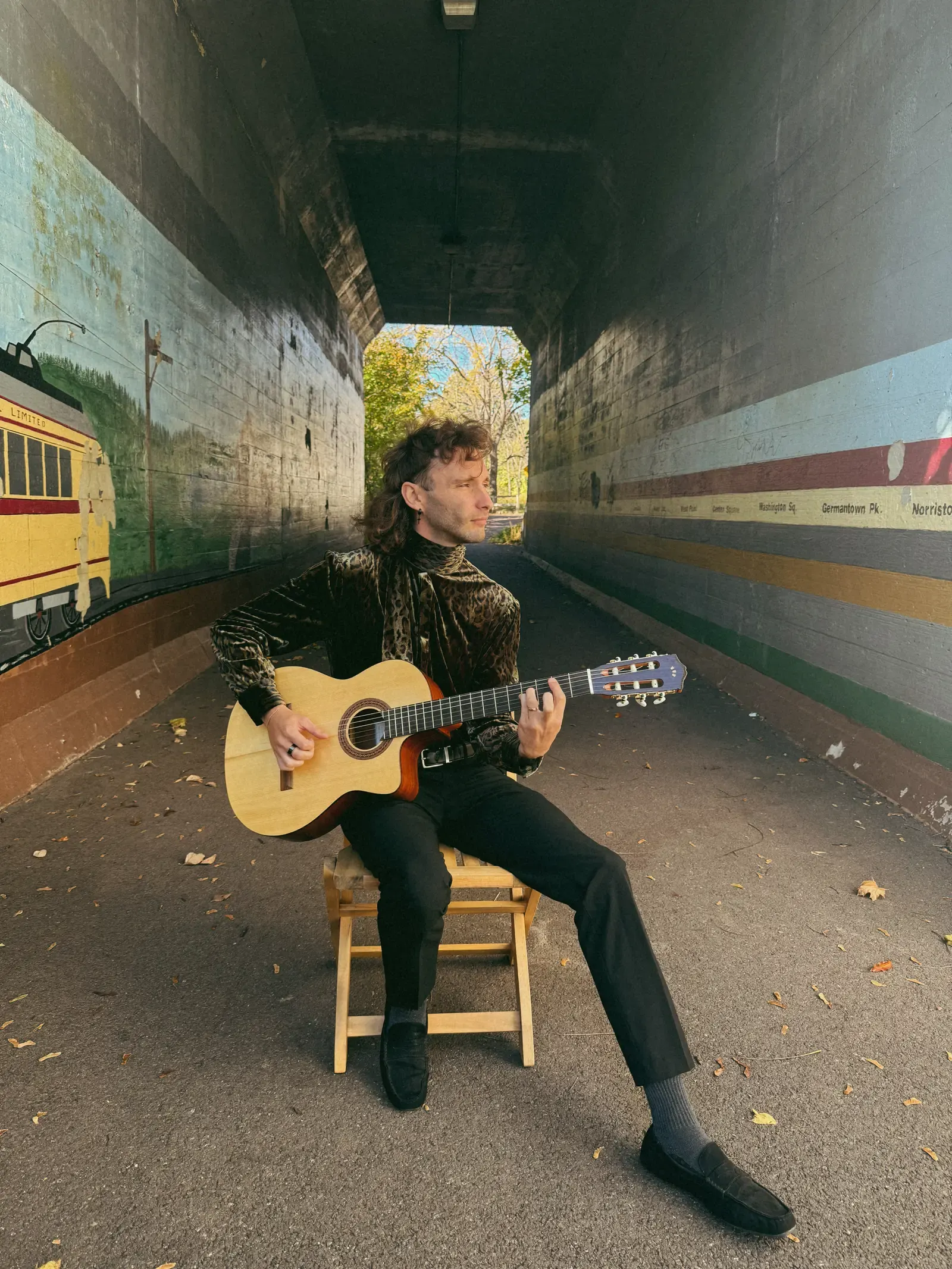 Mathew seated with a pale-wood guitar in a painted passageway.