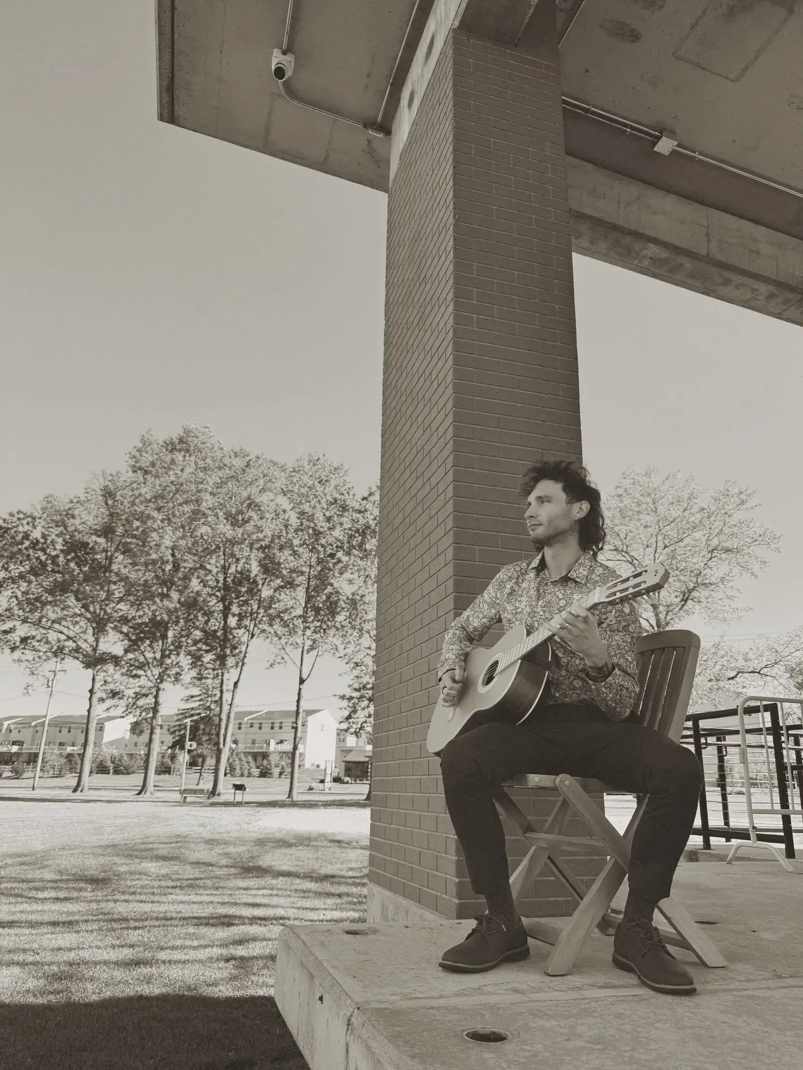 A monochrome portrait of Mathew seated with a guitar beneath a covered walkway.