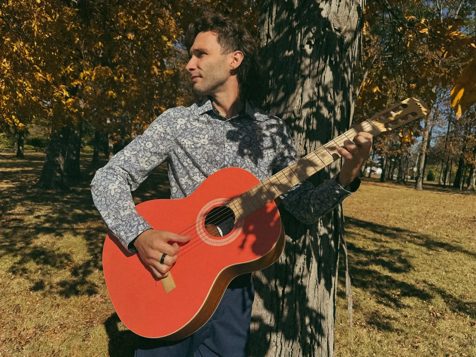Mathew playing his orange-red guitar beneath a tree.